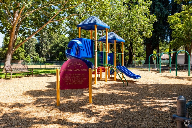 The Willows Oaks Park playground is shaded by large trees.