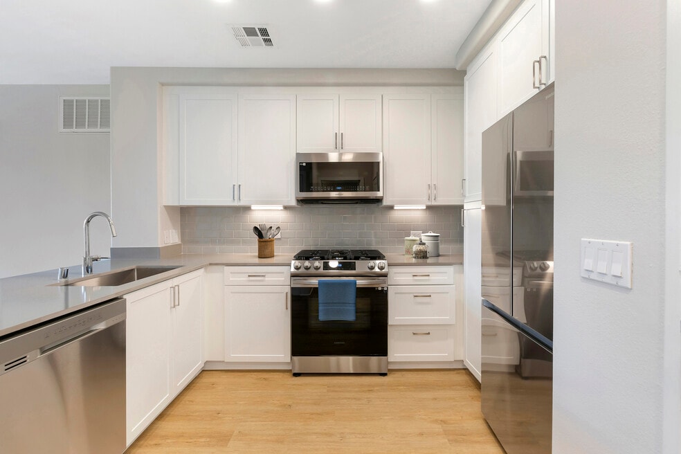 Renovated Package I kitchen with stainless steel appliances, grey quartz countertops, white cabinetry, grey tile backsplash, and hard surface flooring