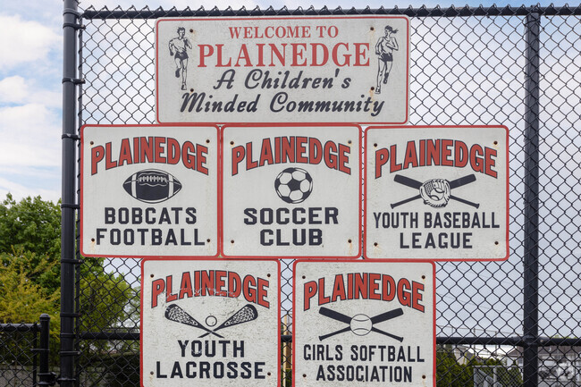 A welcome to Plainedge, NY sign hangs on a ballfield fence greeting all the spectators.