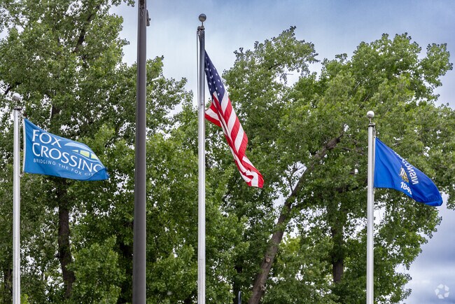 The Fox Crossing, United States, and Wisconsin flags wave together beneath the trees.