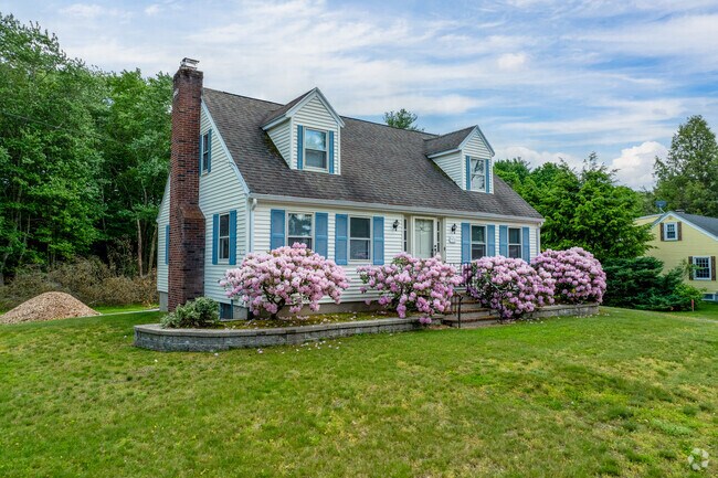 A cape cod style home in Maynard has colorful flowers in front and a large green lawn.