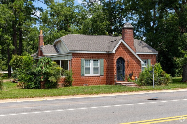 A charming brick home with green shutters and a gabled roof sits along a tree-lined road.