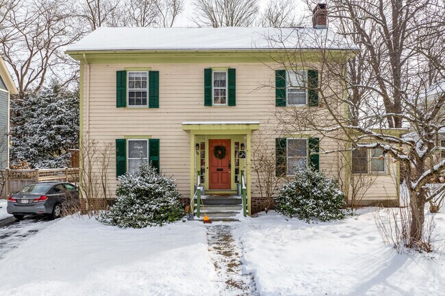 A colonial revival style home in Northampton has a green shutters, yellow portico and beige clapboard siding.