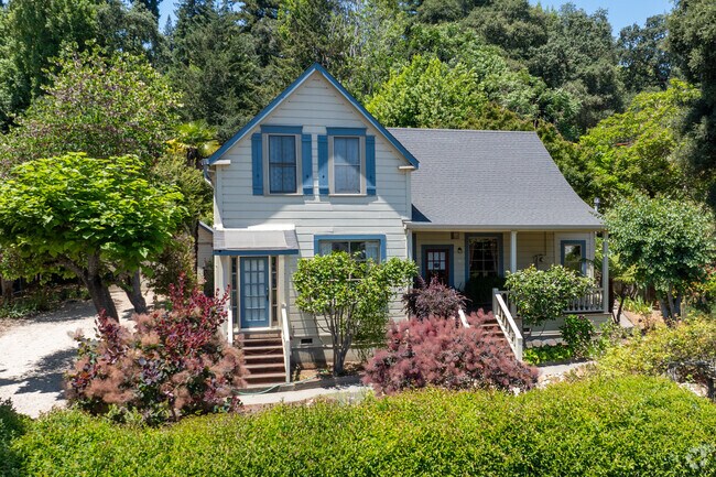 This Folk Victorian home in Felton features shuttered gables, a covered porch, and cottage-style charm nestled in a colorful, tree-lined garden setting.
