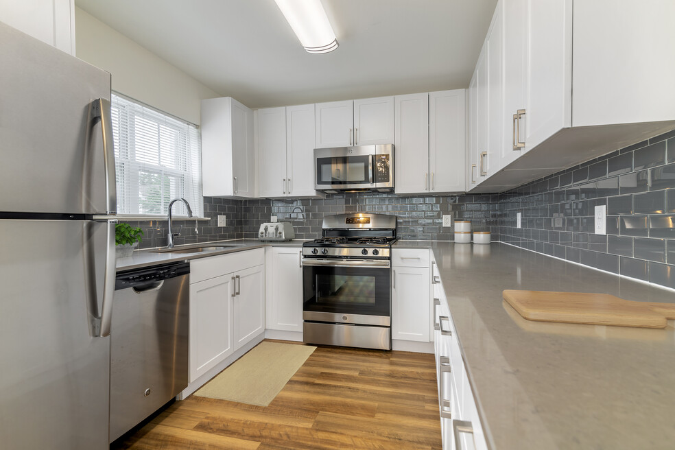Renovated Package II kitchen with white cabinetry, grey quartz countertops, grey tile backsplash, stainless steel appliances, and hard surface plank flooring throughout common areas. Soft-close cabinets and microwaves available in select homes
