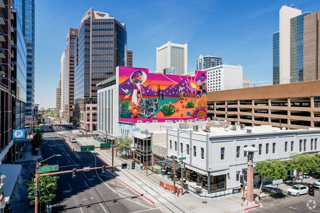 A vibrant mural burst with color in the heart of Downtown Phoenix, where modern skyscrapers rise behind historic buildings and light rail lines weave through the energetic city core.