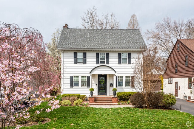 A white Colonial style home sits on a beautiful cherry blossom lined street in Millburn.