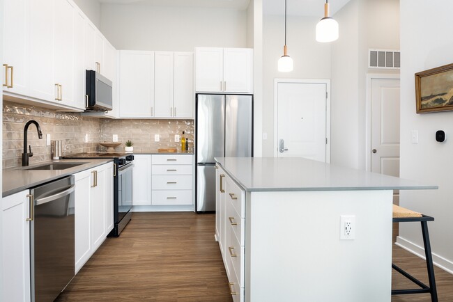 Upgrade Collection kitchen with white cabinetry, beige tile backsplash, grey quartz countertops, pendant lighting, and hard surface vinyl plank flooring (Representative photo)