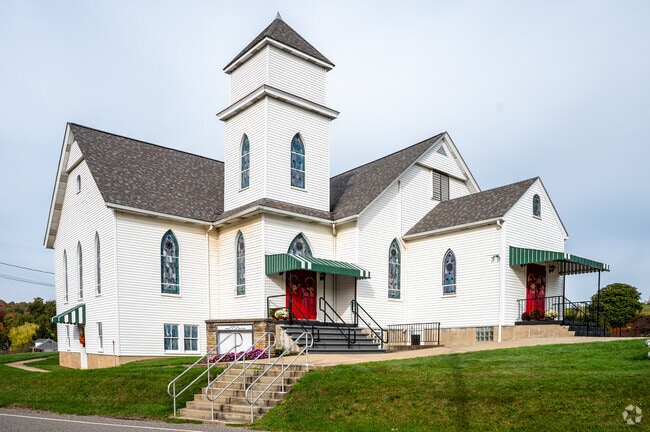 Historic white church in Manor Township Armstrong, featuring a tall central steeple, arched stained-glass windows, and vibrant red doors accented by green awnings.