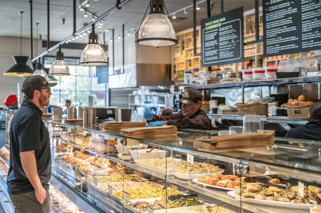 The deli counter at Woodlands Market is busy with customers ordering custom sandwiches.