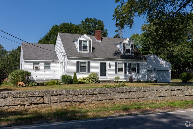 A white Cape Cod home with black shutters sits behind a stone wall and summer perennials.