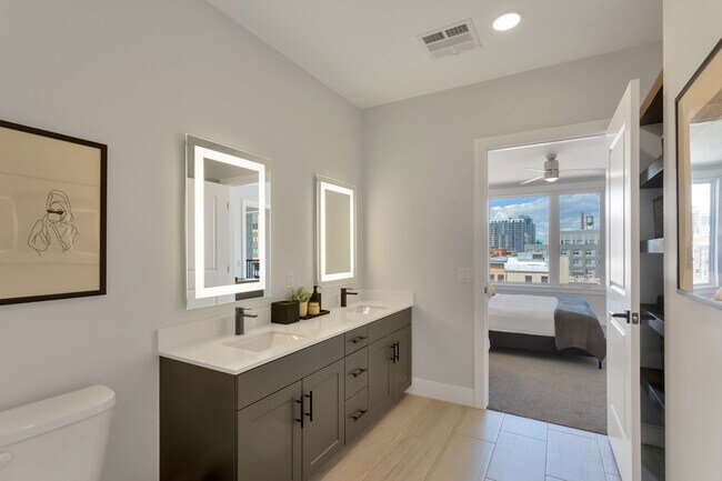 Modern bathroom with dual sinks and illuminated mirrors, flowing into a bright bedroom with large windows offering city views.