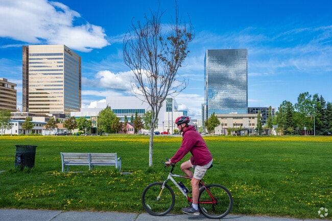 A man rides his bike around Delaney Park with downtown Anchorage behind him