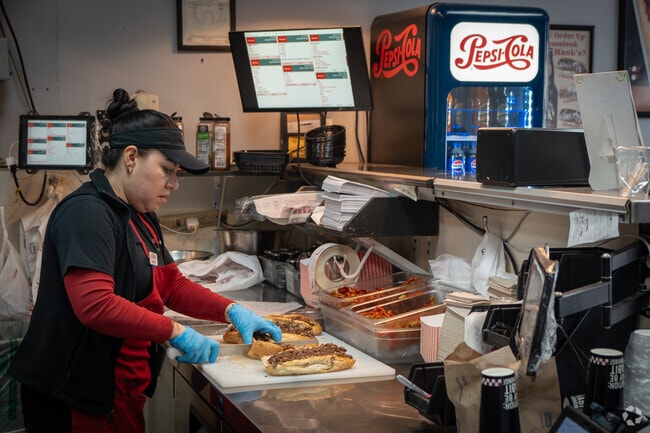 A lady at The Cheese Steak Shop preparing fresh cheesesteaks for hungry customers.
