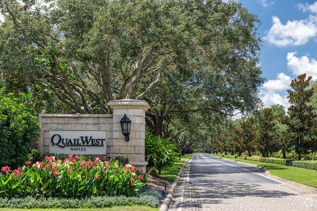 The entrance to Quail West features a long driveway line with large arching trees.